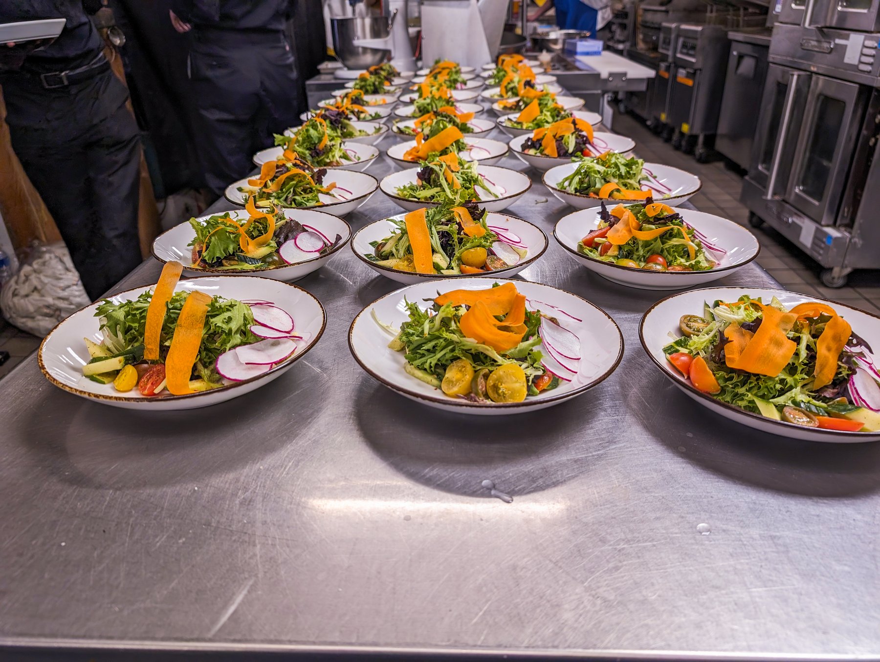 Rows of plated salads with greens, radish, and carrot ribbons prepared on a stainless steel kitchen counter